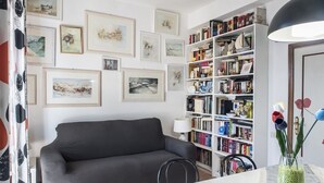 Ett vardagsrum med en grå soffa, en bokhylla full av böcker och dekorativa föremål, och en vägg prydd med inramade bilder.