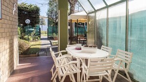 En täckt uteplats med ett runt bord och vita stolar, en glasveranda och utsikt över omgivningen.