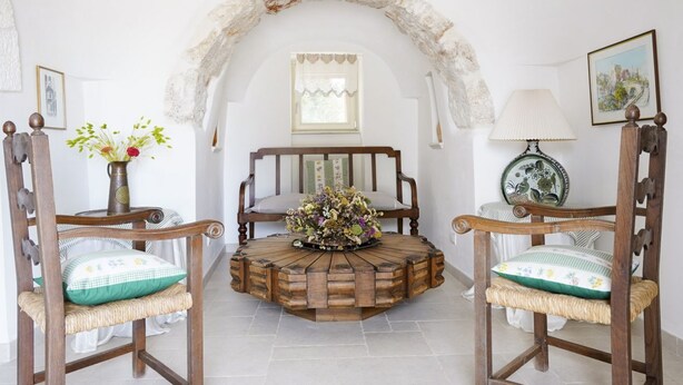 A room with a round wooden coffee table, two wooden chairs with cushions, a vase with flowers, a lamp, and a framed picture on the wall.