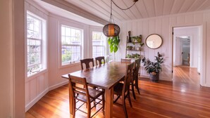 Un comedor con mesa y sillas de madera, ventanas amplias y una lámpara colgante.