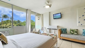 Quarto de hotel com uma cama grande, um sofá pequeno, uma televisão e uma varanda com vista para palmeiras.