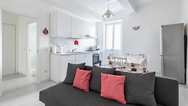 Ett modernt kök med vita skåp, en grå soffa med röda kuddar och en matplats med ett bord och stolar.