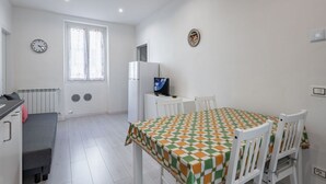 A small, bright room with a dining table, chairs, a small refrigerator, a television, and a window with curtains.