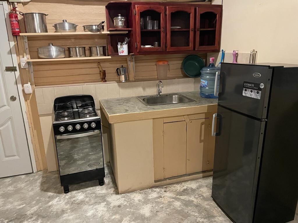 A compact kitchen with a stove, sink, and refrigerator. There are wooden cabinets above the counter and a fire extinguisher on the wall.