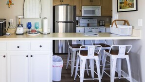 A kitchen with a stainless steel refrigerator, a microwave, a stove, and a sink. There are white bar stools and a white basket on the counter.