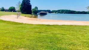 A lakeside view with a sandy beach, a covered structure, and a large tree.