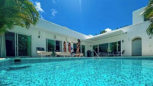 En swimmingpool med flisegulv, en hvid bygning med store vinduer og en klar blå himmel.