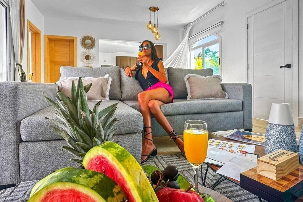 Un salon moderne avec un canapé gris, une table en verre sur laquelle se trouve une boisson, et des fruits tropicaux.