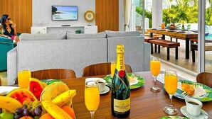 Ein modernes Wohnzimmer mit einer Couch, einem Esstisch gedeckt mit Früchten, einer Champagnerflasche und einem Fernseher an der Wand.