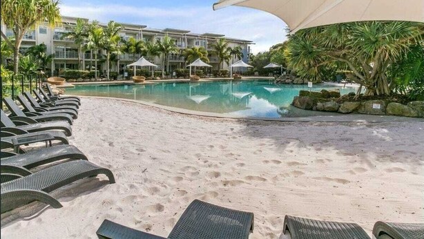 A resort pool area with lounge chairs, a large umbrella, and a building in the background. - The Sand Resort Kingscliff - Stylish & Spacious Apt 8121/22 - Holiday Management Co. (Tweed)