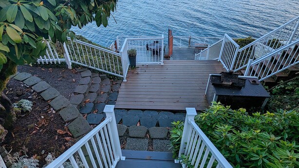 Una terraza de madera con barandas blancas que conduce a un muelle sobre un lago, rodeado de vegetación.