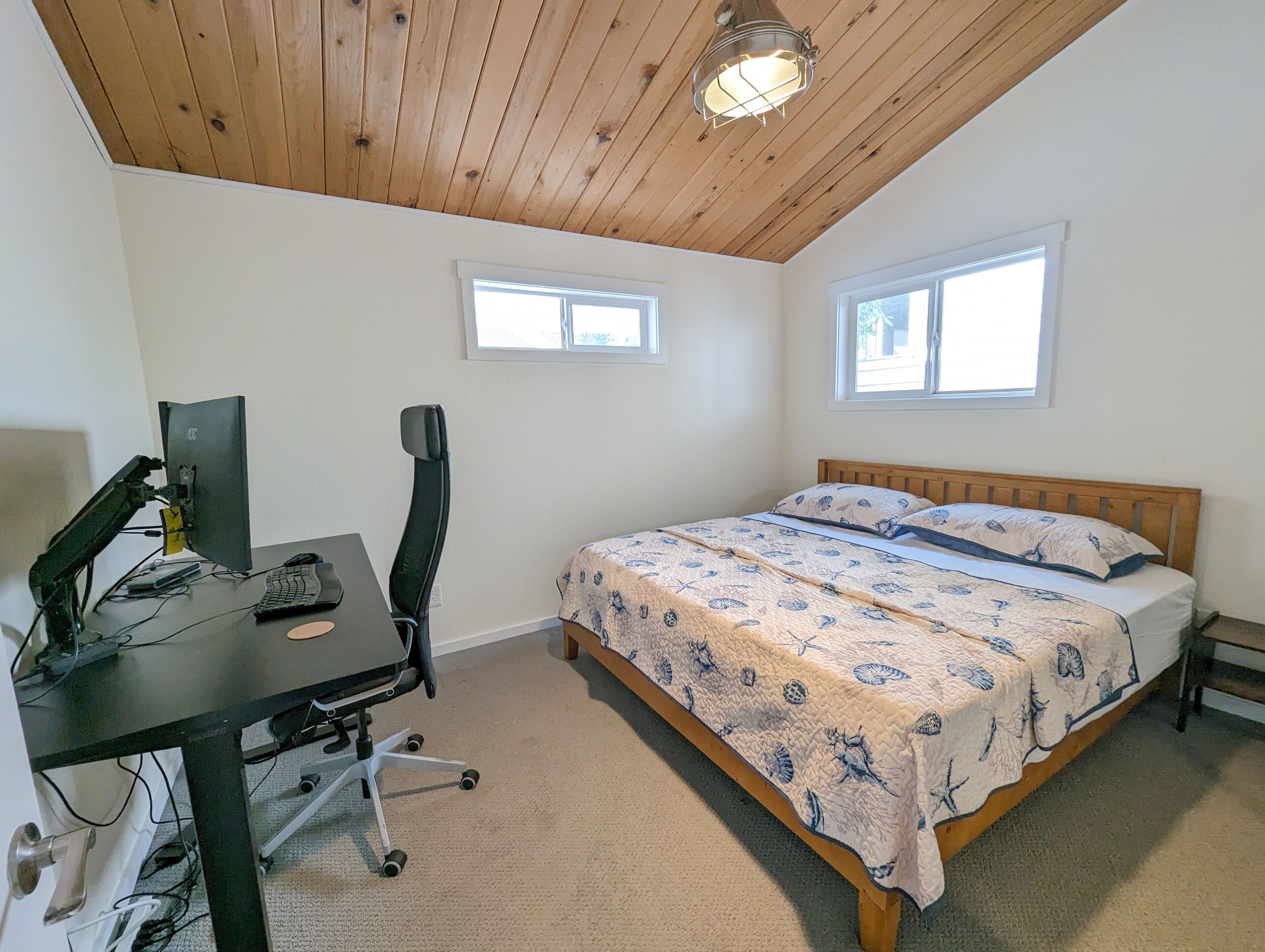 Une chambre avec un plafond en bois, un bureau avec un ordinateur, une chaise, un lit recouvert d’une couvre-lit à motifs et deux fenêtres.