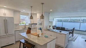 Una cocina moderna con armarios blancos, una isla central con taburetes, iluminación colgante y vistas a un lago a través de amplios ventanales.