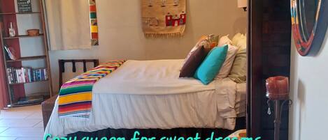 A cozy bedroom with a bed, colorful pillows, a rug, a bookshelf, and a wall tapestry.