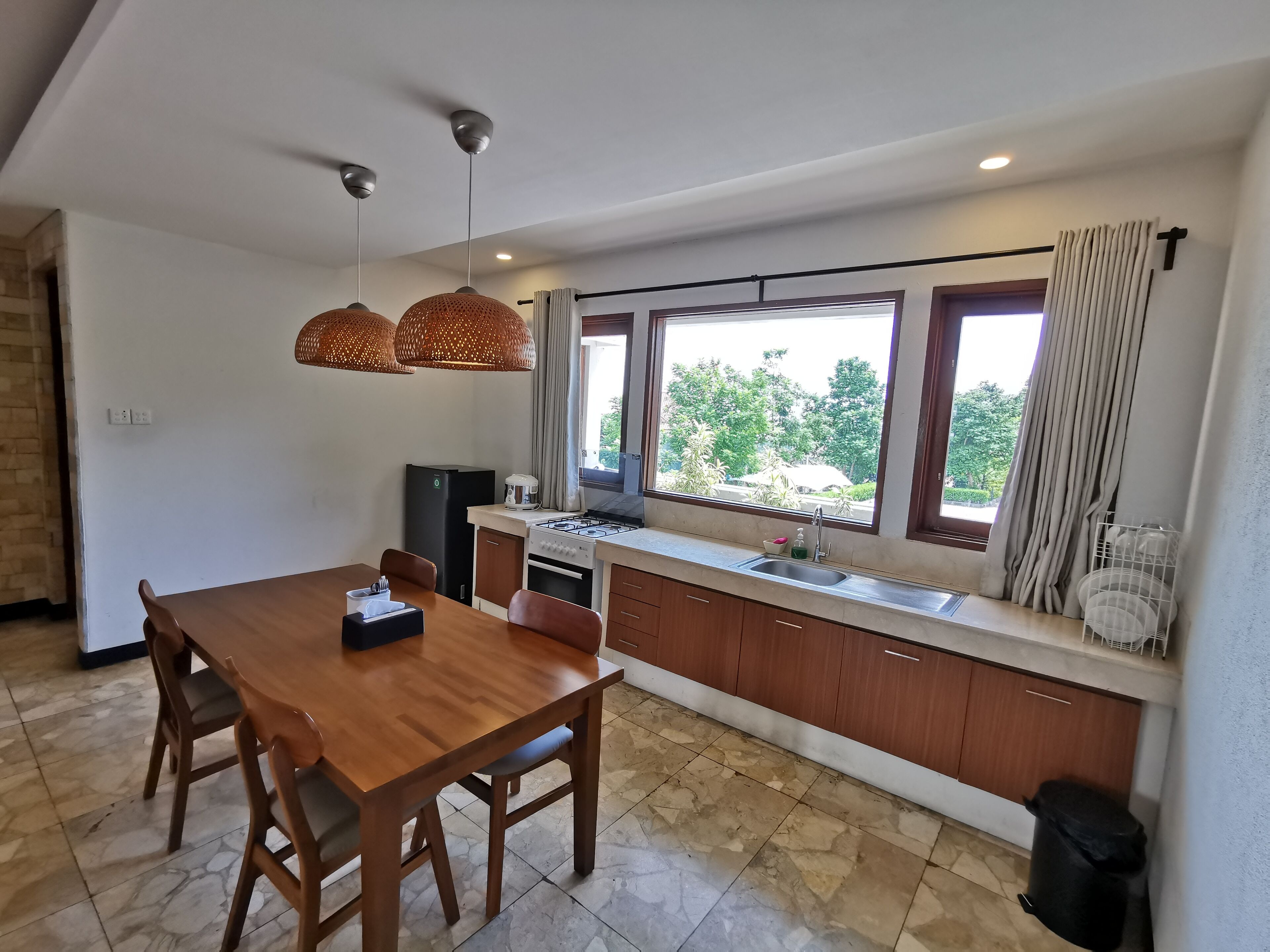 A modern kitchen with a dining area, large windows, and a view of greenery outside.