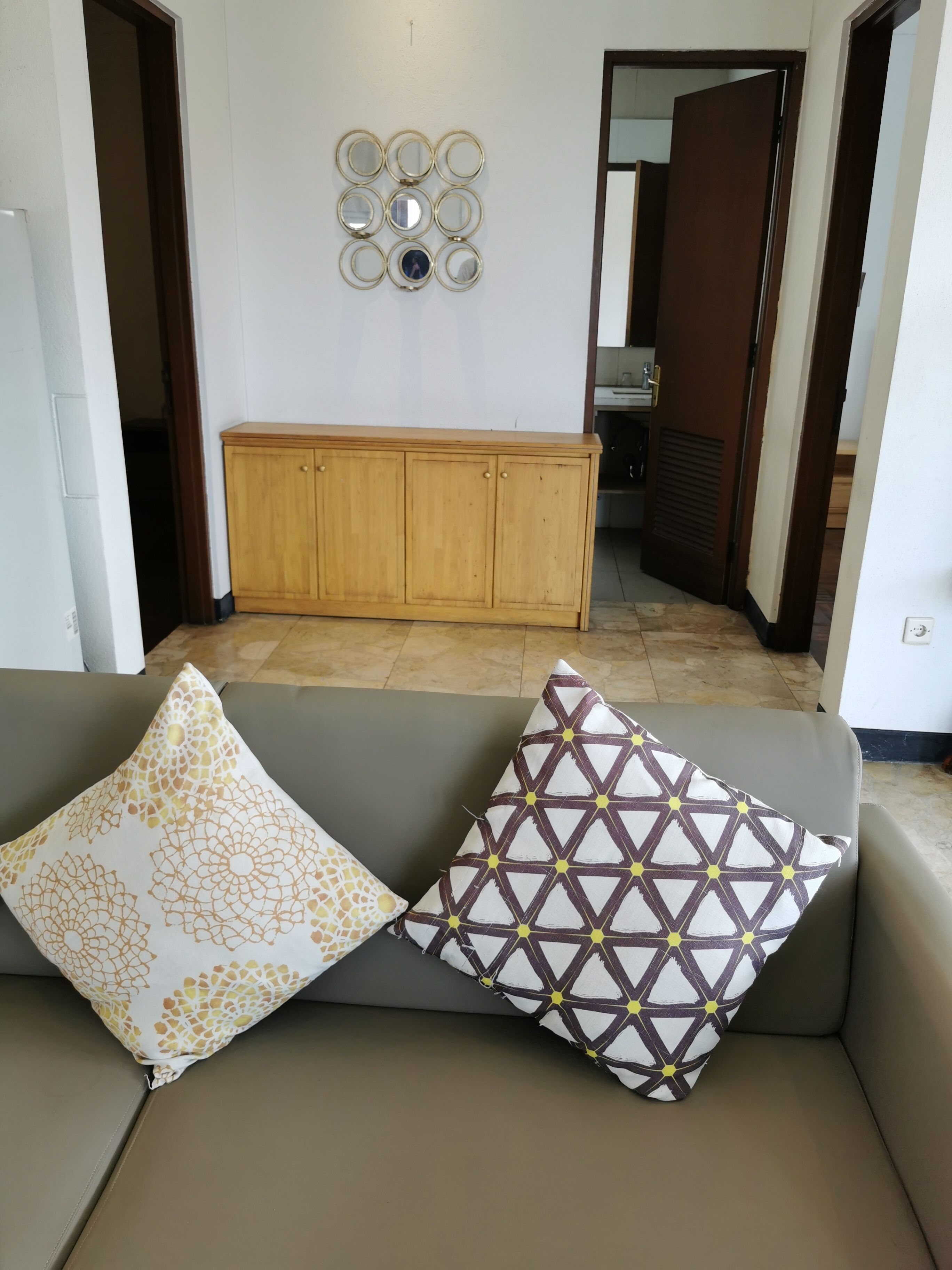 Un salón con un sofá, dos cojines estampados, un mueble de madera y un espejo instalado en la pared.