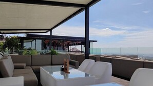 A modern outdoor lounge area with a glass coffee table, white sofas, and a view of the cityscape.