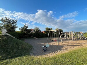 Un parque infantil con un sube y baja, columpios y un tobogán, rodeado de césped y árboles.