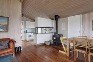 A cozy interior with a wooden floor, a dining area with a table and chairs, a kitchen with white cabinets, and a black wood-burning stove. - Beautiful sunsets 100 meters from the beach (Brenderup)