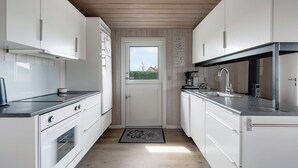 A narrow kitchen with white cabinets, a built-in oven, and a window with a view of greenery. - Beautiful sunsets 100 meters from the beach (Brenderup)