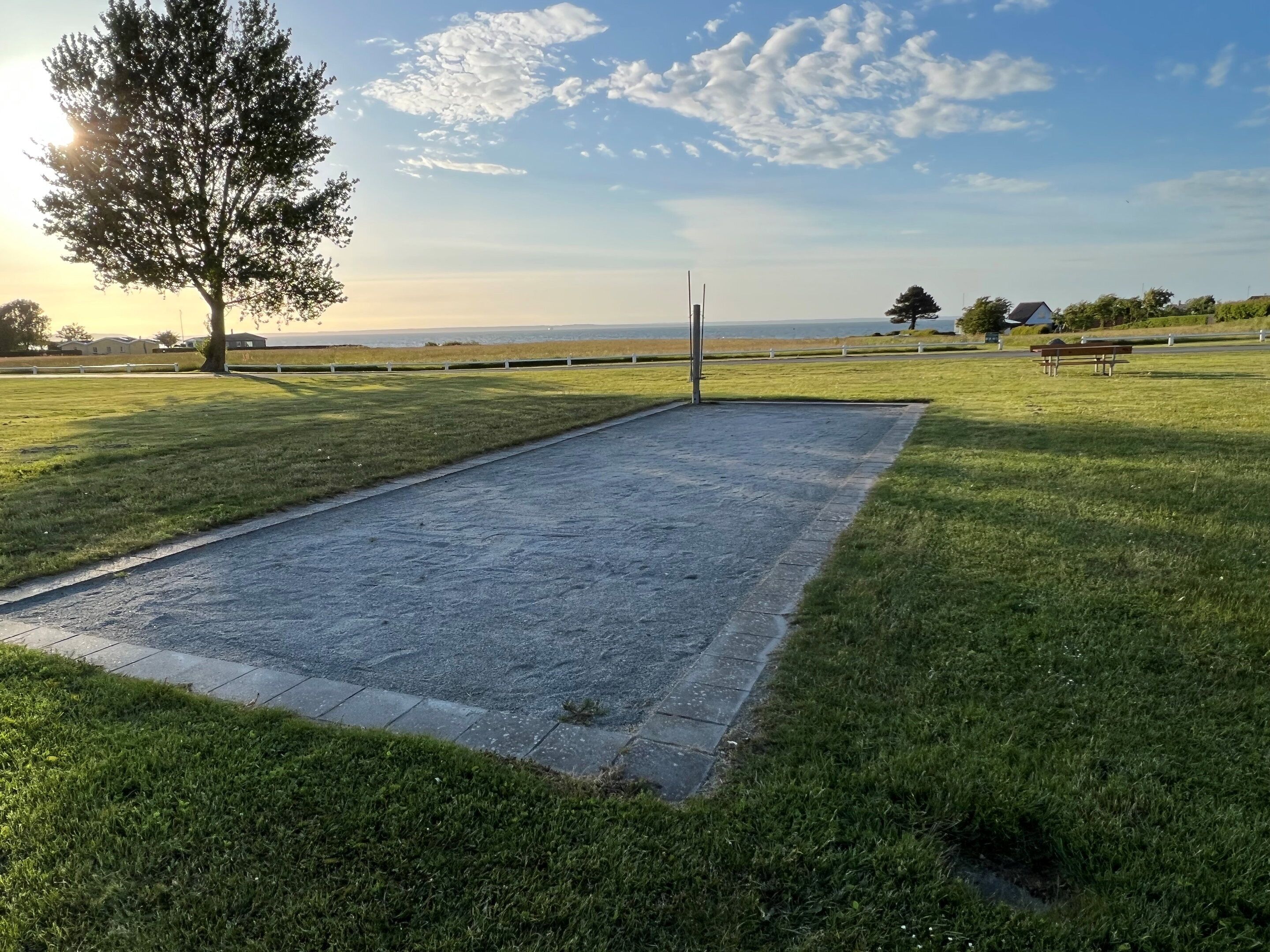 A rectangular paved area with grass on either side, a tree, and a distant view of a body of water.