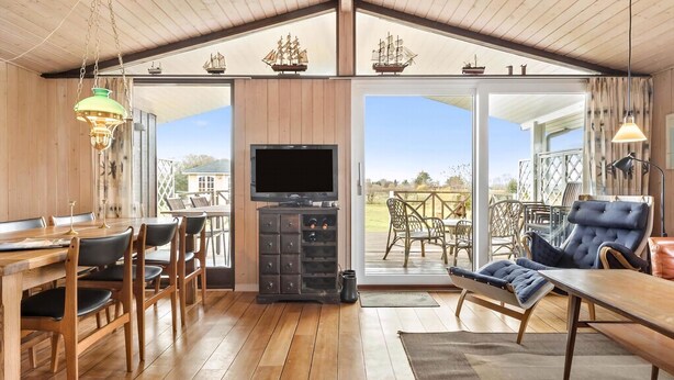 A wooden interior with a TV, dining table, chairs, and a balcony with wicker furniture. - Beautiful sunsets 100 meters from the beach (Brenderup)