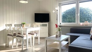 A modern living room with a dining area, a sofa, a TV, and a large window with a view of greenery. - DA25/1 - Family-friendly, comfortable vacation apartment with garden use - House (Cuxhaven)