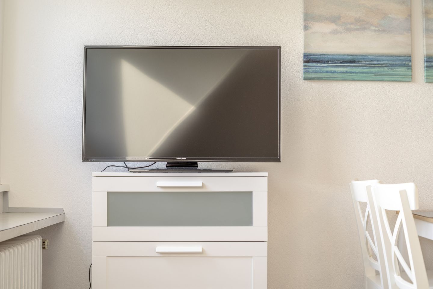 Ein Flachbildfernseher an einem weißen Schrank mit zwei Schubladen, ein weißer Stuhl und ein Gemälde an der Wand.