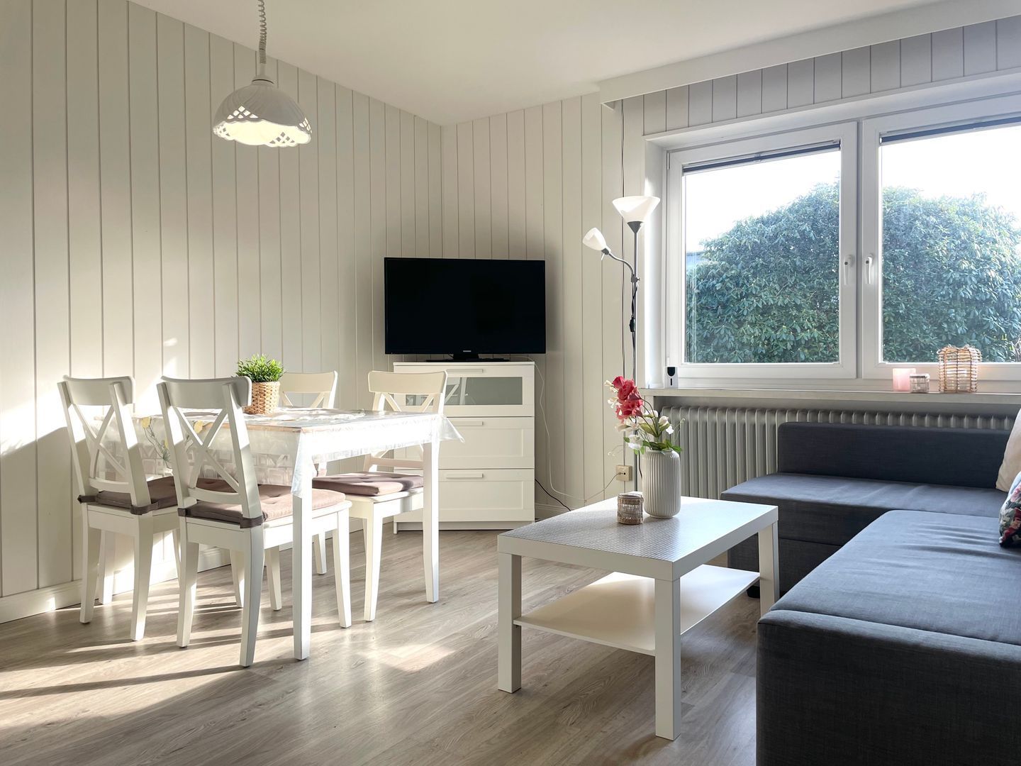 Un salon moderne comprenant un coin repas, un canapé, un téléviseur et une grande fenêtre donnant sur un paysage verdoyant.