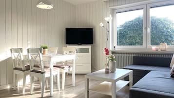 Ein modernes Wohnzimmer mit Essbereich, einer Couch, einem Fernseher und einem großen Fenster mit Blick auf Grünflächen.