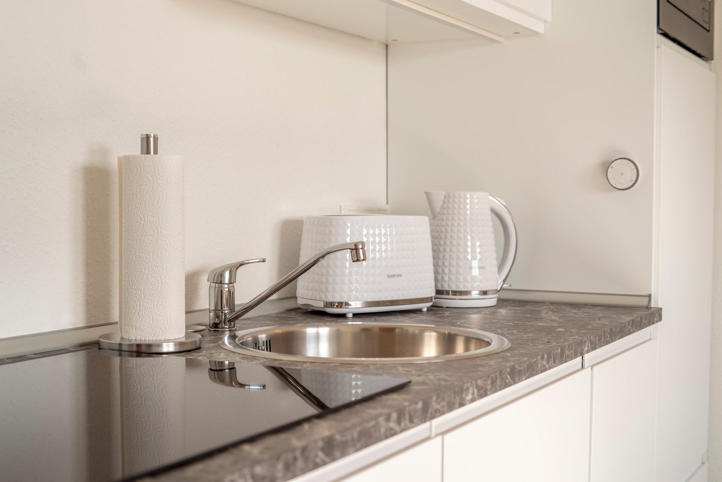 Een keuken met een spoelbak van roestvrij staal, een moderne kraan, een witte waterkoker en een rol keukenpapier.