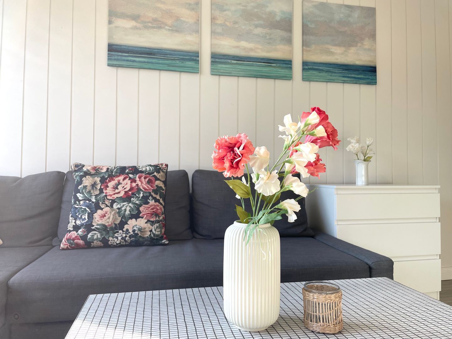 Ein Wohnzimmer mit einer Couch, einem floralen Kissen, einer Vase mit Blumen, einem kleinen Tisch und einer Wand mit Gemälden.