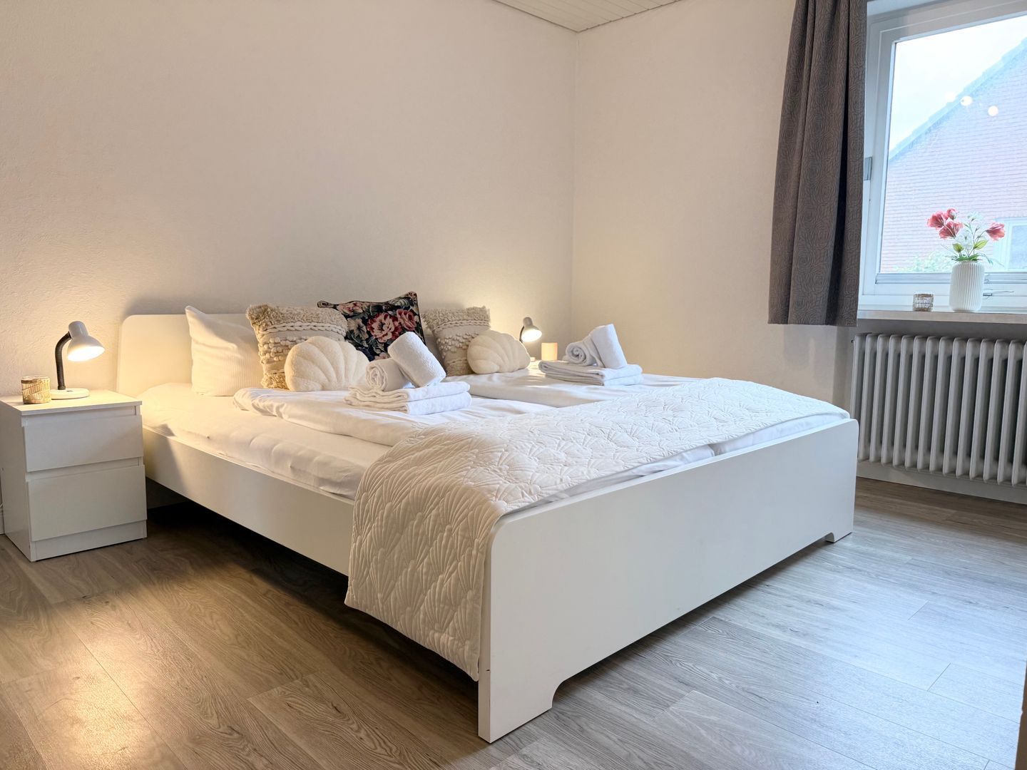 Un lit bien fait, avec du linge de lit et des serviettes blanches, une lampe de chevet et une fenêtre donnant sur un jardin fleuri.