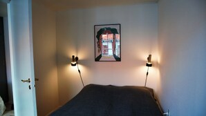 A room with a bed, two wall-mounted reading lamps, and a framed picture on the wall. - Cozy apartment in old farm (Svaneke)