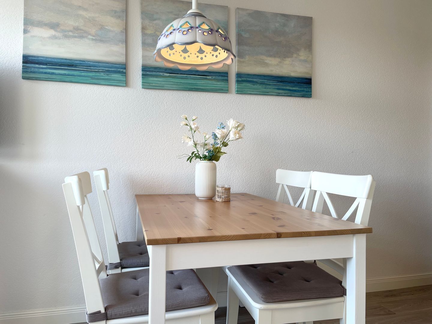 Un coin repas avec une table en bois, des chaises blanches, un vase de fleurs et une suspension.