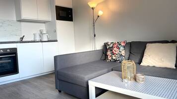 Eine moderne Küche mit weißen Schränken, einem eingebauten Ofen und einer Kaffeemaschine. Im Wohnbereich befindet sich eine graue Couch mit floralen Kissen und ein weißer Tisch mit einem gewebten Korb und einem Glas.