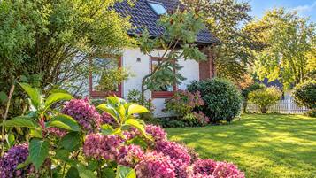 Ein Haus mit geneigtem Dach, umgeben von üppigem Grün und blühenden Pflanzen.