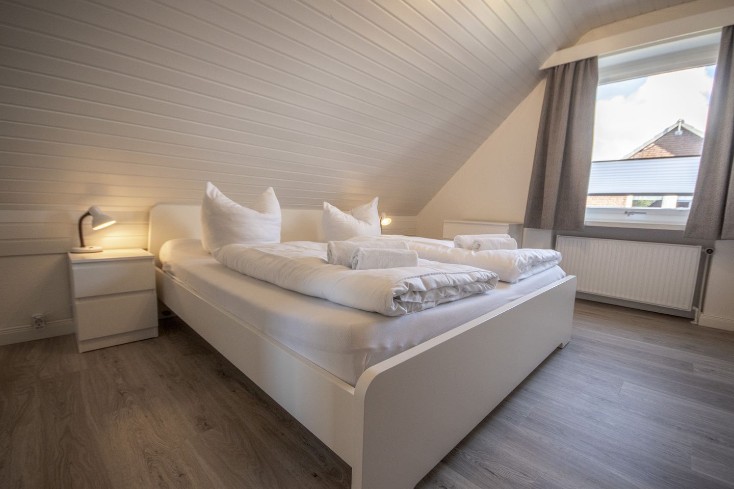 Ein Schlafzimmer mit schrägen Wänden, einem Bett mit weißer Bettwäsche, einem Nachttisch mit Lampe und einem Fenster mit Vorhängen.