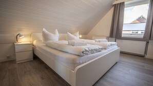 A bedroom with a sloped ceiling, a bed with white bedding, a nightstand with a lamp, and a window with curtains.