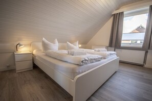 Un dormitorio con techo inclinado, una cama con ropa blanca, una mesita de noche con lámpara y una ventana con cortinas.