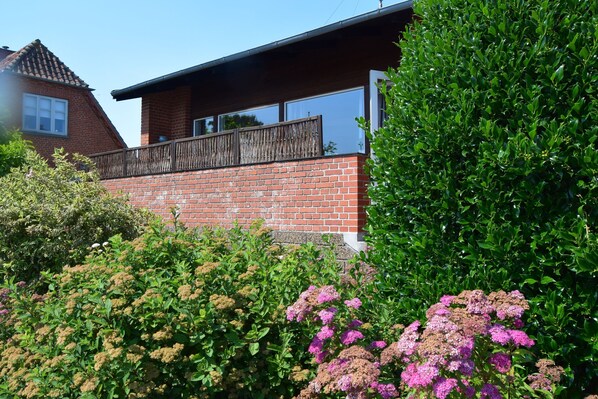 A residential building with a brick facade, a balcony, and dense green shrubbery.