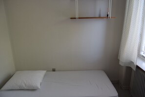 A small, single-bed room with a white mattress, a white pillow, a wooden shelf, and a window with a curtain.