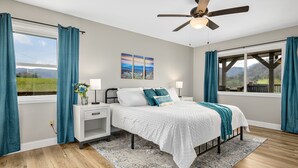 A bedroom with a bed, nightstands, a window with curtains, a ceiling fan, and a view of the outdoors. - NEW Breathtaking Views, Hot tub and Arcade, in a Peaceful Mountain View Retreat (Clyde)