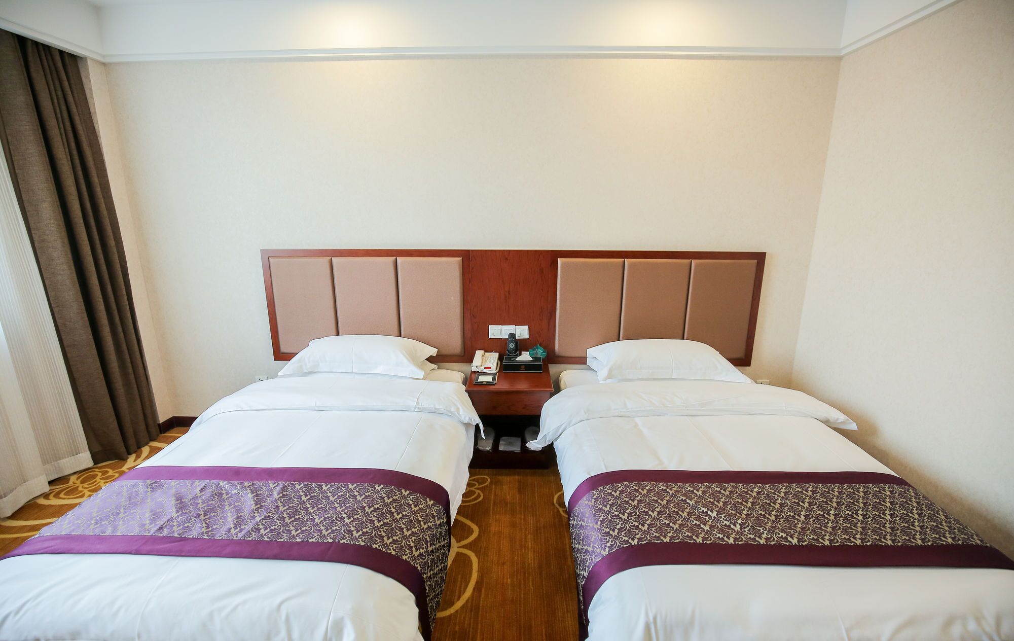 A hotel room with two single beds, a wooden headboard, a nightstand with a phone and a lamp, and a carpeted floor.