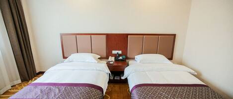 A hotel room with two single beds, a wooden headboard, a nightstand with a phone and a lamp, and a carpeted floor.