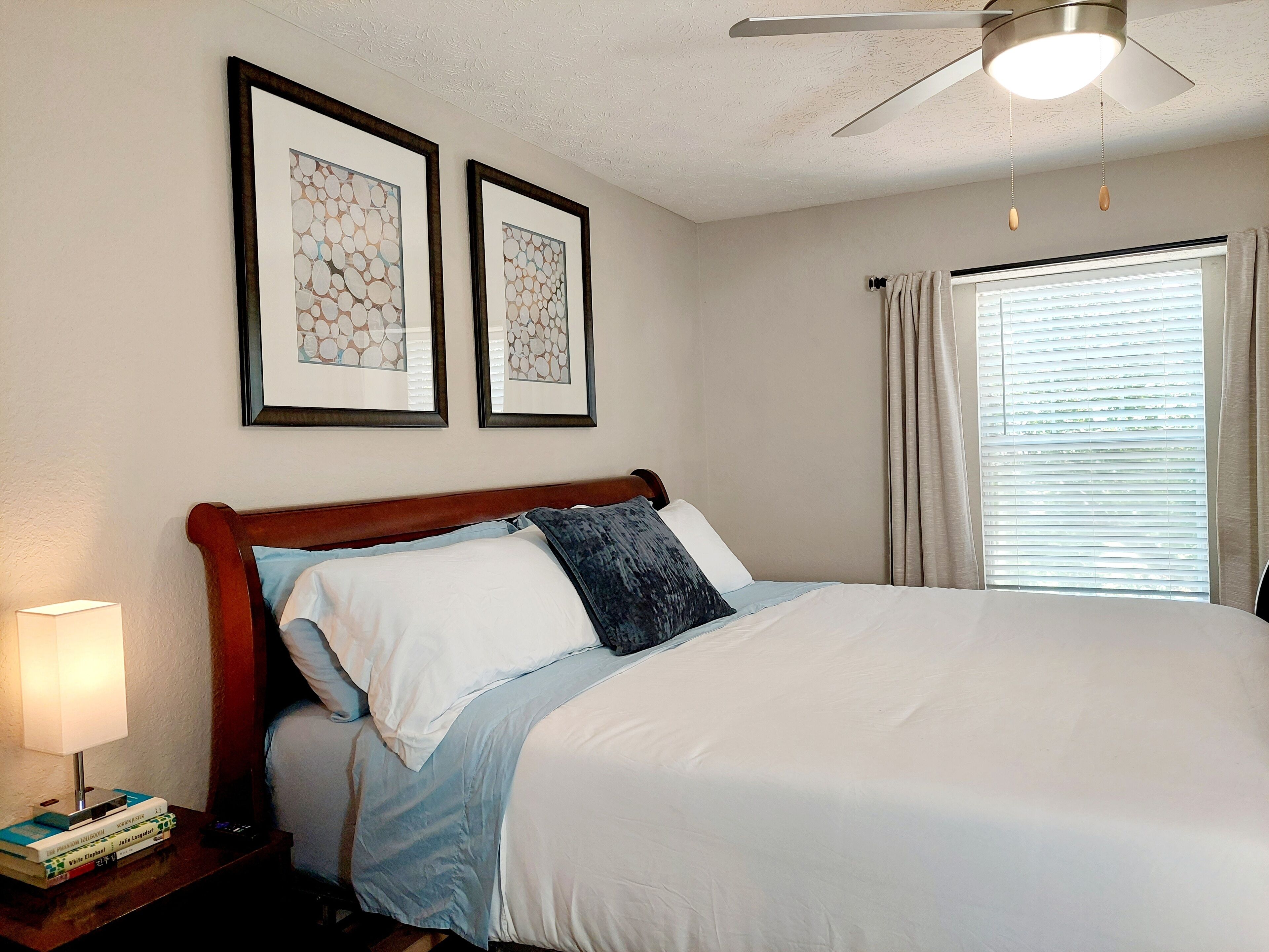 A neatly made bed with a wooden headboard, a bedside table with a lamp and books, and two framed artworks on the wall.