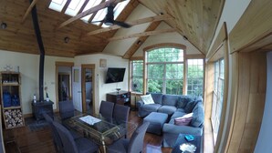 A spacious living area with a high ceiling, wooden beams, a glass coffee table, and a large window offering a view of trees. - Beachfront Guest Cottage in Antigonish (Antigonish)