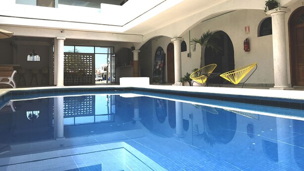 Piscina com superfície de água azul límpida, rodeada por paredes e colunas brancas. Há cadeiras amarelas e um vaso de planta perto da piscina.