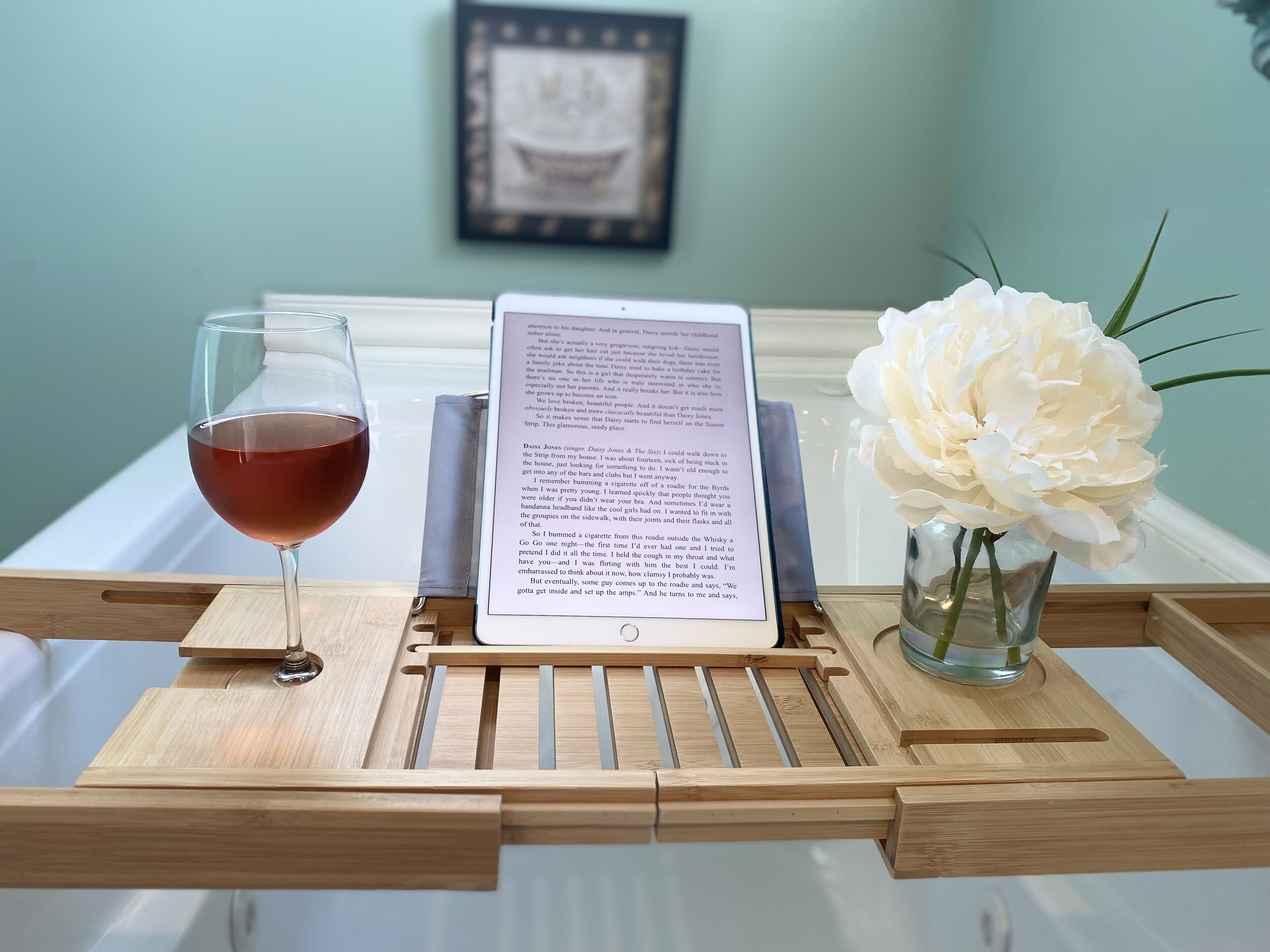 A wooden tray with a glass of red wine, an open book, and a white flower in a glass vase.