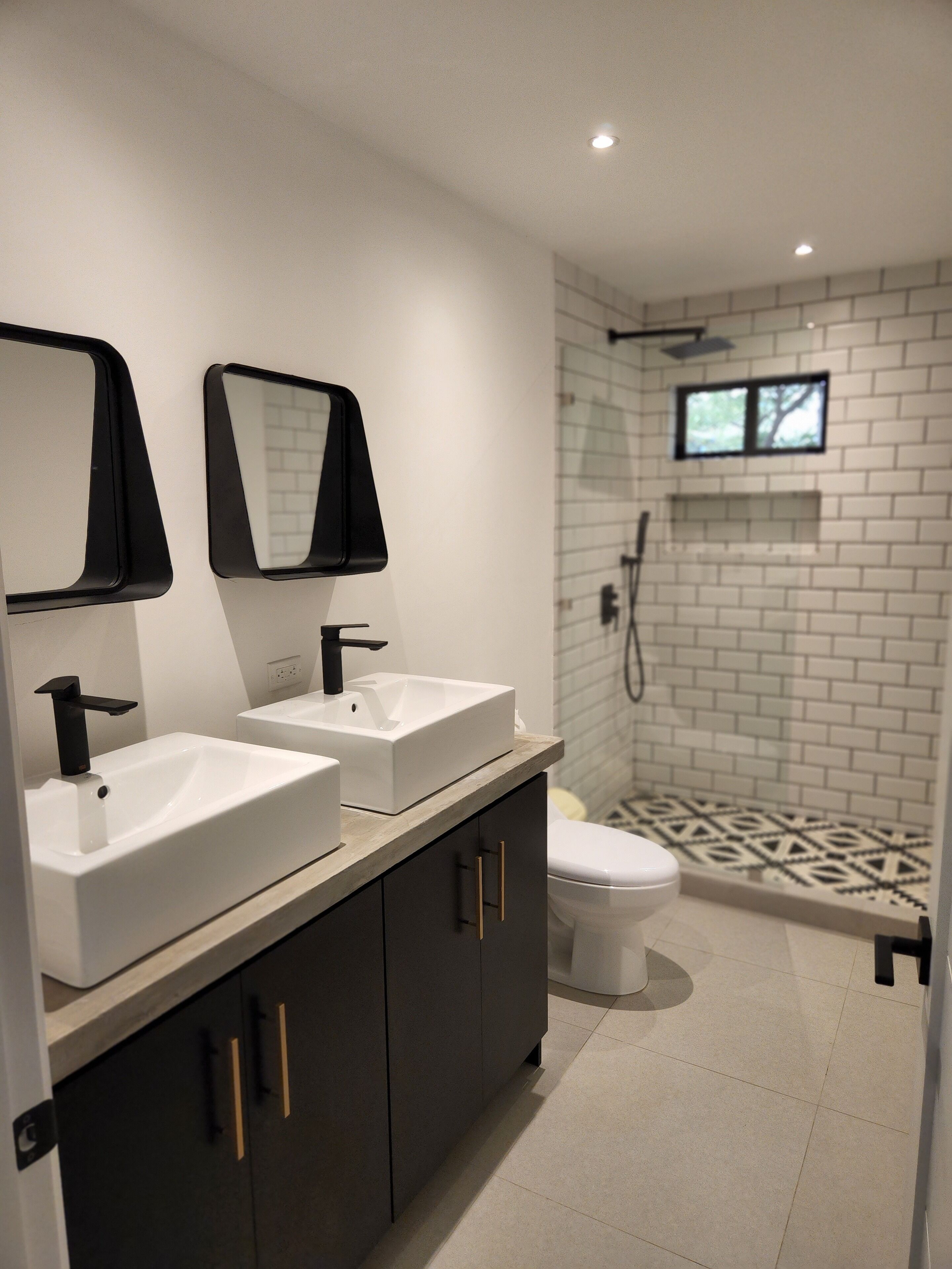 A modern bathroom with two white sinks, black faucets, and a toilet. There is a shower area with a glass door and a small window. The floor has a patterned tile design.